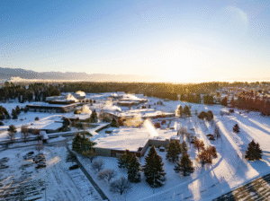 An aerial drone view of the snow-covered Flathead Valley Community College campus in Kalispell at sunrise, with steam rising from buildings and mountains in the distance.