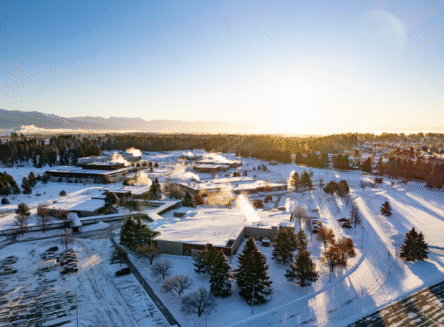 An aerial drone view of the snow-covered Flathead Valley Community College campus in Kalispell at sunrise, with steam rising from buildings and mountains in the distance.