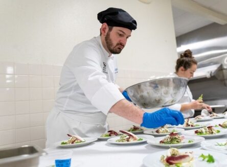 Culinary students in white chef uniforms and hats work in a professional kitchen, preparing food and garnishing plates.