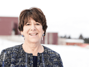 A portrait of Flathead Valley Community College President Jane Karas smiling outdoors in a snowy setting, wearing a blue and black patterned jacket and drop earrings.