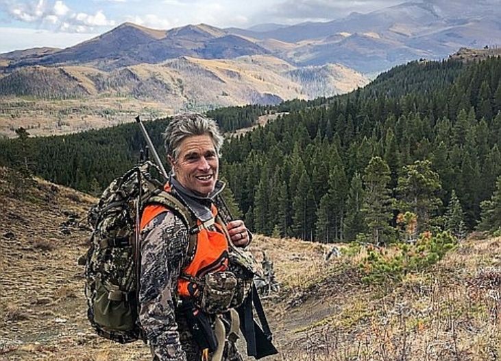 Franz Ingelfinger wearing hunting gear and a large backpack standing on a mountain hillside with a forest and peaks in the background.