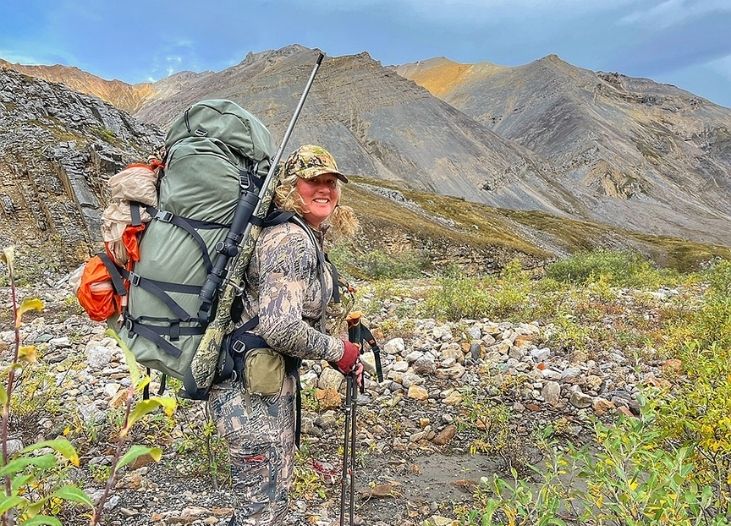 Rachel Schmidt wearing camouflage hunting gear and a large hiking pack standing in a rocky mountain landscape.
