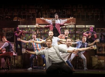 he Choir of Man performers on a theater stage set as a vintage pub, with cast members holding beer steins and playing instruments like the violin and guitar.