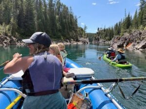 A group of people in a blue raft and a green kayak floating down a calm river surrounded by tall evergreen trees and rocky cliffs.