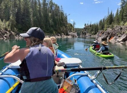 A group of people in a blue raft and a green kayak floating down a calm river surrounded by tall evergreen trees and rocky cliffs.