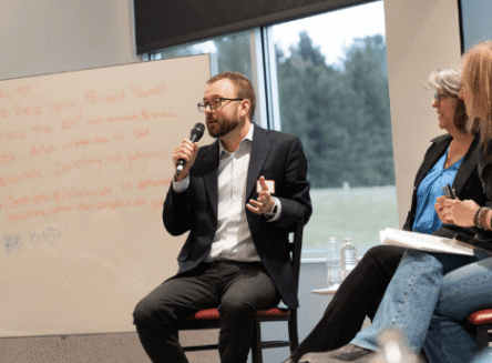 A man in a dark blazer and glasses speaking into a microphone while seated on a panel next to two women, with a whiteboard of Guidelines visible in the background.