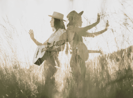 A warm-toned, sun-drenched photograph of the musical duo Rising Appalachia, featuring two women standing back-to-back in a field of tall grass while holding an acoustic guitar and a banjo.