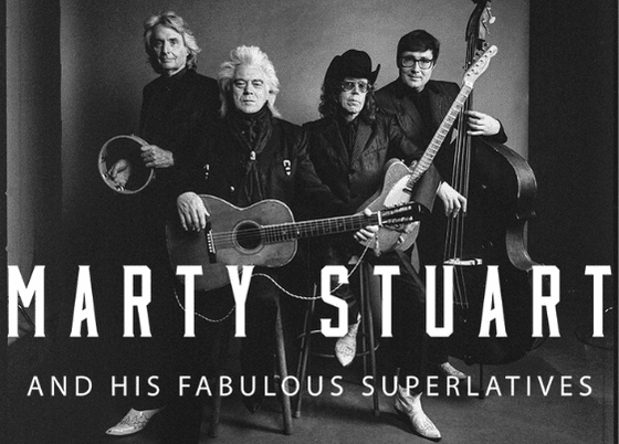 A group portrait of the four members of Marty Stuart & His Fabulous Superlatives standing in a line. All are wearing sharp, matching all-black outfits with fitted jackets and Western-style boots. They are clean-cut with signature hairstyles and pose confidently with their electric guitars and a bass.